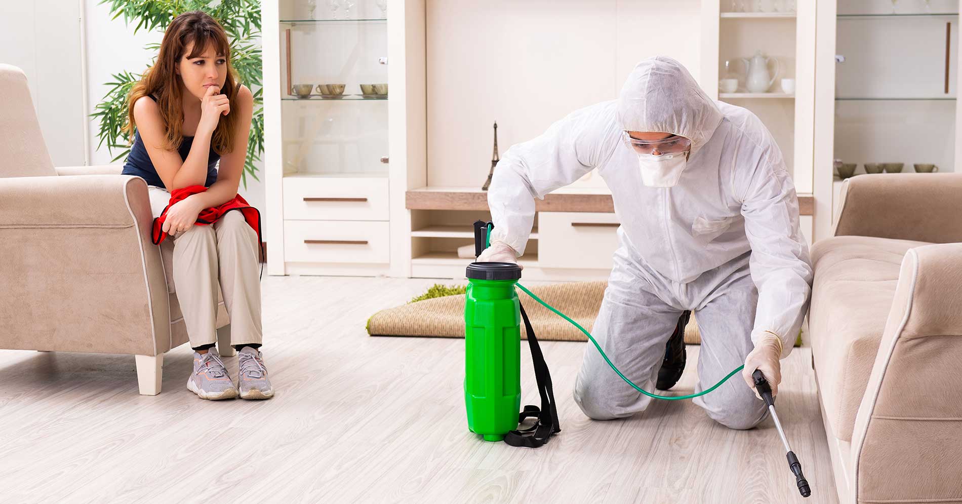 Person in einem Schutzanzug sprüht Desinfektionsmittel unter ein Sofa